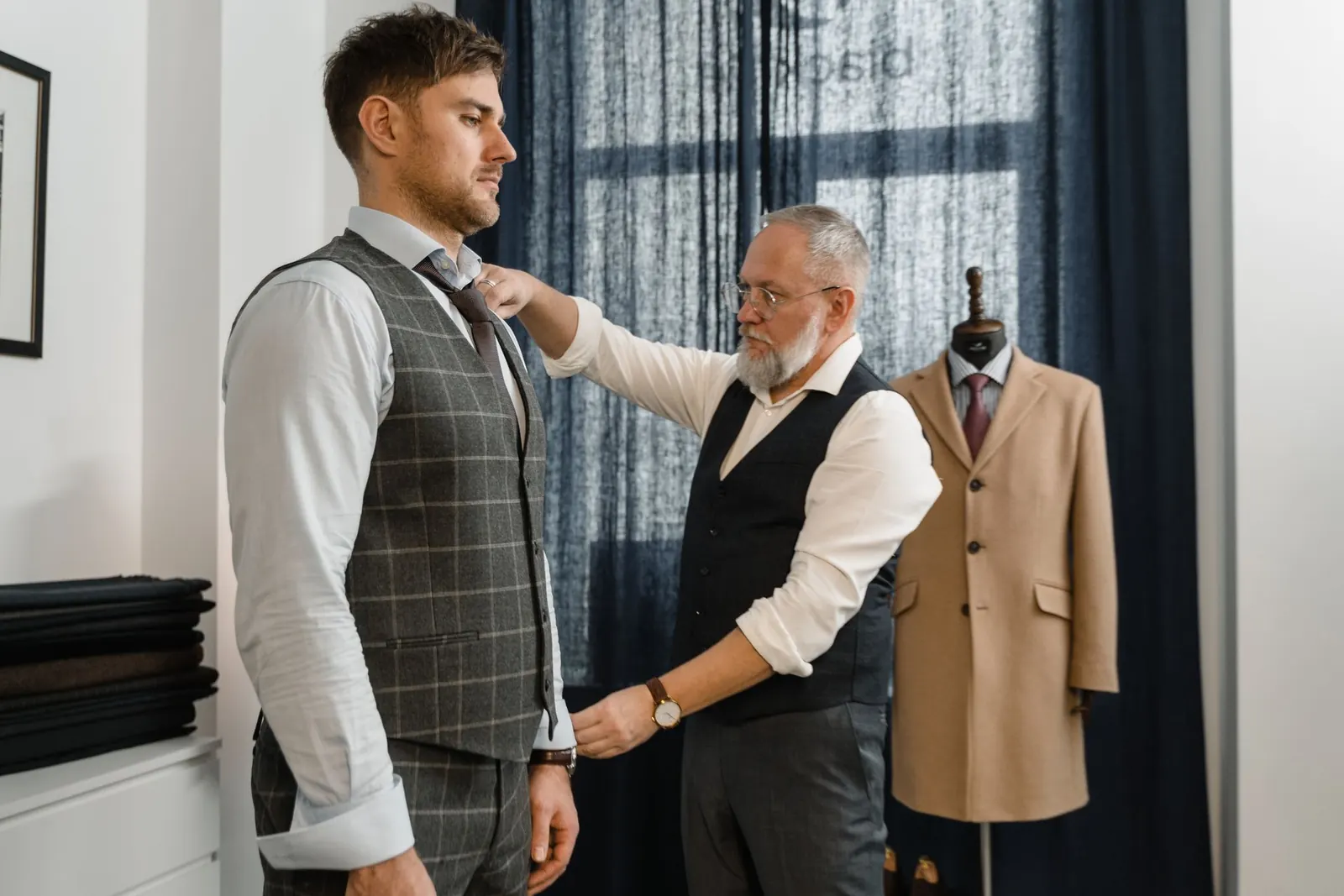 A tailor in a vest pinning the collar of a suited male client during a fitting